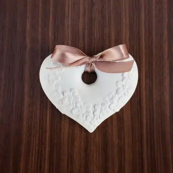 Pingente decorativo em forma de coração, com padrão floral em relevo e laço acetinado castanho.