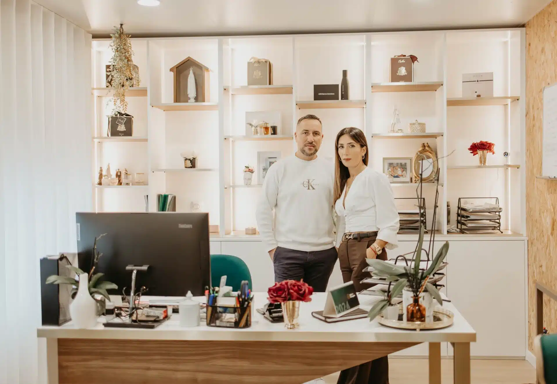 Duas pessoas na receção de um escritório, junto a uma estante iluminada com objetos decorativos.